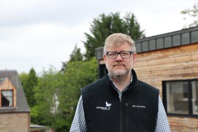 Person wearing glasses in front of a wooden hut