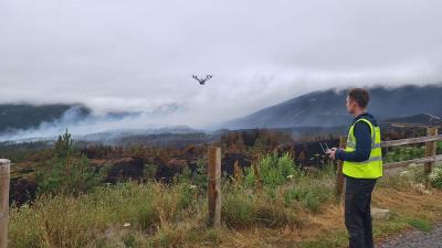 A thermal drone pilot in a yellow hi-vis vest looks out across the scene of the Glenmore wildfire