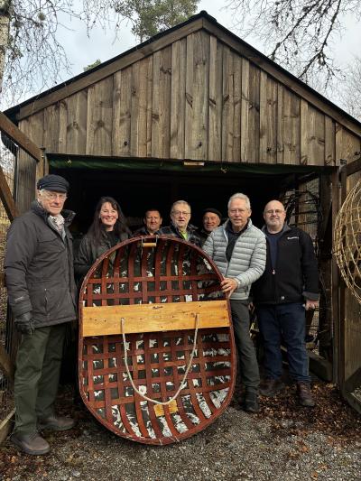 group of seven people holding a traditional handmade boat in front of a shed