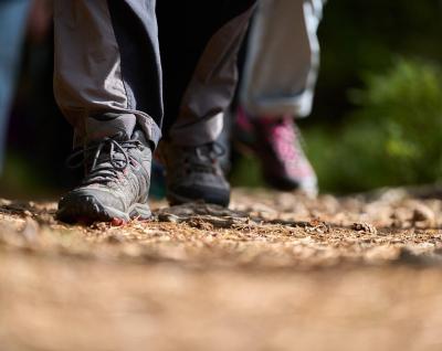 Close up photo of the feet of a person wearing grey walking boots. Walking on a brown forest track with another person's feet visible behind them