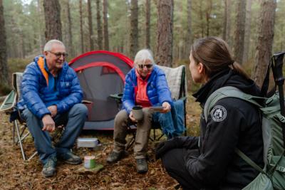 Two people sitting on camp chairs in a forest in front of their tent, talking to a countryside ranger