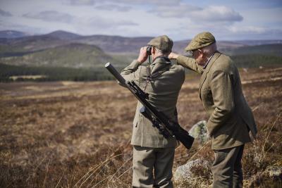 Two men in tweed jackets and flat caps observe the landscape with binoculars. One of the men has a gun slung over his shoulder.