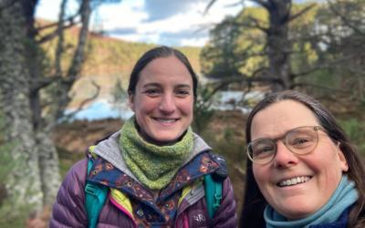 Two women smiling taking a selfie in the outdoors with trees and a loch in the background.