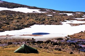 Wild camping in a small tent, next to a small lochan with snow in the background.